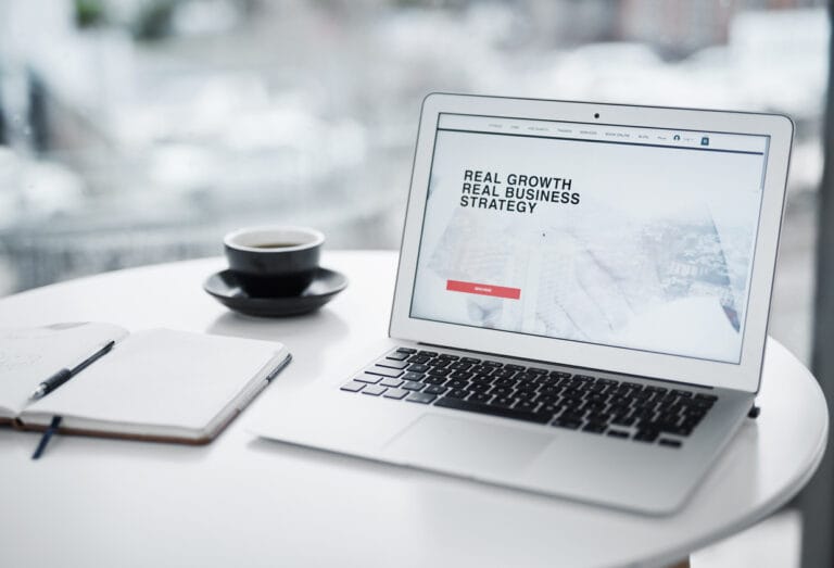 A laptop displaying text Real Growth Real Business Strategy on screen, and a cup of coffee and a notebook on a table