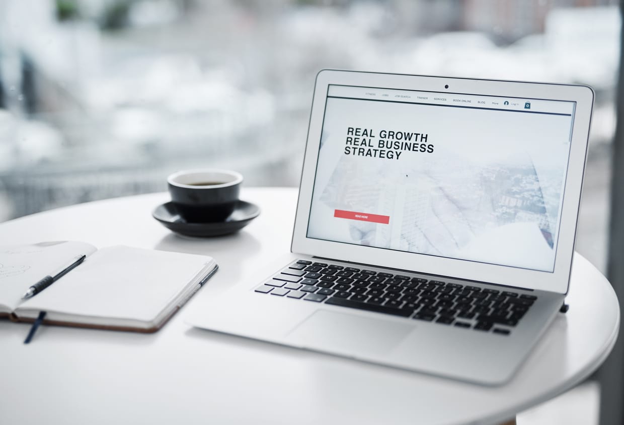 A laptop displaying text Real Growth Real Business Strategy on screen, and a cup of coffee and a notebook on a table
