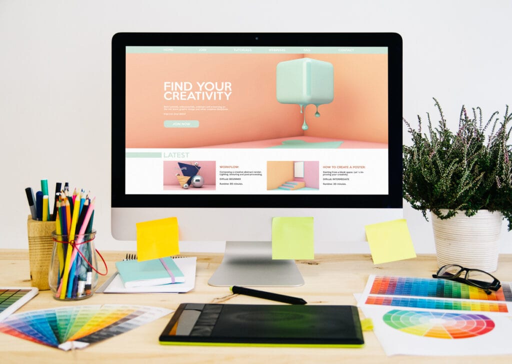 A webpage is shown on a computer monitor placed on a table with stationery, design elements, and a graphic tablet.