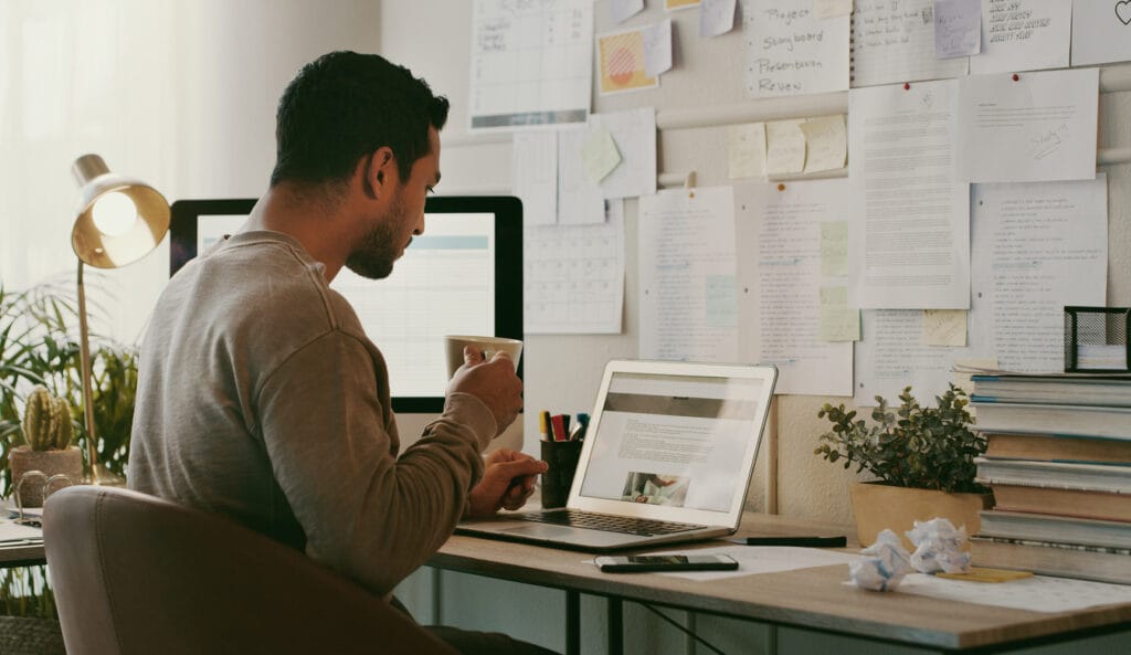 Shot of a young businessman sitting in his home office and drinking coffee while using his laptop