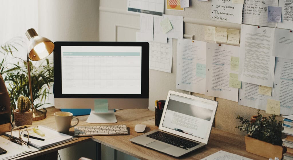 Streamline content workflow: A laptop and computer monitor on a corner office desk with documents pinned on the wall